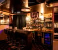A stylish bar area with a long bar counter, several high stools, shelves filled with bottles, a large mirror, a small table with a flower arrangement, and decorative lighting.