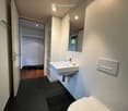 Modern bathroom with white walls, black tiled floor, white sink, mirror, and toilet. An open closet with wooden flooring is visible through the doorway.