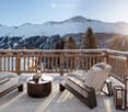 A wooden balcony with a scenic mountain view, featuring a sofa, a round table, and two chairs with fur throws.