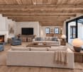 Cozy living room with exposed wooden beams, fireplace, large windows offering a view, and comfortable seating including sofas, armchairs, and ottomans.