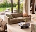 Modern living room with large windows, a beige sectional sofa, coffee table, armchairs, and a TV. The room features a view of a well-maintained garden and patio area with additional seating.