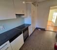 Modern kitchen with white cabinets, black countertop, and built-in oven and dishwasher. The floor is carpeted, and a doorway leads to another room with wooden flooring.