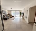 Modern living room with a grey sectional sofa, coffee table, side table, and artwork on the walls. The space features large windows with sheer curtains and a view of a balcony area.