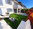 Well-maintained garden with artificial grass, white sofa, glass coffee table, sun loungers, and colorful foliage.