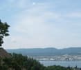 Scenic outdoor view showing a lake, distant mountains, and a town across the water, with trees and a roof visible in the foreground.