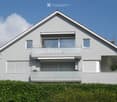 Modern two-story house with grey siding, large windows, and a balcony on the upper floor.