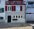 Two-story house with red shutters, white windows, and a red balcony railing. The ground floor has a door and several windows, while the upper floor features additional windows and a small balcony. The house is situated on a cobblestone street.
