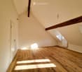 Empty attic room with wooden flooring, slanted ceilings, and exposed wooden beams. Natural light streams in from a skylight window.