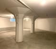 Empty underground space with concrete floor, white walls, and support pillars. Features a small window and fluorescent lighting.