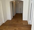 Empty hallway with light wooden flooring, white walls, and a single hanging light fixture. Doors visible at the end of the hallway.