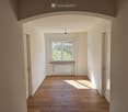 Empty hallway with wooden flooring, white walls, and a window at the end. Natural light streams in, casting shadows on the floor.