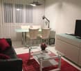 Modern living room with a black leather couch, red pillows, a glass coffee table, and a dining set with a glass table and chairs. A TV is on a cabinet, and a lamp is on the side table. A red rug is on the floor.