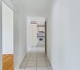 Empty hallway with white tiles, white walls, wooden floor in one part, kitchen in the background
