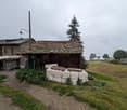 A small wooden building with a stone roof and a larger wooden building in the background on a cloudy day. Several trees and plants are in the area. The road leads to the buildings.