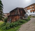 old wooden house, located at the end of a driveway, beside a modern building, surrounded by vegetation