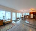 Open layout, large windows, hardwood floors, dining table and chairs, kitchen with cabinets and countertop, white appliances, rug.