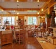 This image shows a cozy, rustic-style restaurant or cafe interior. The space features wooden beams and paneling, giving it a warm and inviting atmosphere. There are several dining tables with chairs, as well as shelves and decor items along the walls. A f