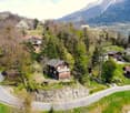 Aerial view of a mountainous, forested area with a small village nestled among the trees. The landscape features winding roads, scattered houses and chalets, and a backdrop of snow-capped mountains.