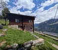Rustic wooden cabin in a mountainous landscape, with a grassy yard and a solar panel visible. The cabin has a balcony or terrace overlooking the scenic mountain views.