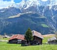 The image shows a picturesque mountain landscape with snow-capped peaks in the background. In the foreground, there are several traditional wooden cabins or chalets surrounded by a lush green meadow. The cabins have red tiled roofs and are nestled among t
