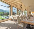 Spacious dining area with large windows offering a scenic mountain view, wooden ceiling beams, pendant lighting fixtures, wooden dining tables and chairs, and a cozy, rustic ambiance.