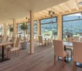 Spacious dining area with large windows offering scenic mountain views, wooden beams and columns, wooden tables and chairs, pendant lighting fixtures, and a cozy, rustic atmosphere.