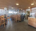 The image shows the interior of a cafe or restaurant. The space has a rustic, wooden decor with a wooden ceiling and walls. There is a long bar counter with stools, and shelves behind the bar displaying various items. The kitchen area is visible in the ba