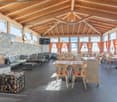 Large open-concept dining area with wooden beamed ceiling, stone fireplace, comfortable seating, and wooden tables and chairs. The space has large windows providing a scenic view of the snowy outdoor landscape.