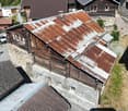 This image shows a traditional alpine-style wooden house with a stone foundation. The roof is made of rusty metal sheets, and the exterior walls are constructed with wooden logs. The house appears to be situated in a mountainous, forested area with other