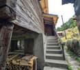 Rustic wooden cabin with a raised ground floor, stone steps leading up to the entrance, and a stack of firewood visible outside.