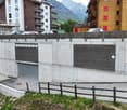 The image shows a multi-story residential building with a concrete and wooden exterior. There is a garage door visible on the ground level, indicating the presence of a garage. The building is surrounded by a wooden fence and is situated in a mountainous