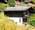 Wooden chalet house, gray roof, wooden balconies, white lower walls, tall grass and trees in front