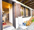 Outdoor view of a wooden house, open entrance, wooden door, tiled floor, wooden table, wooden shelves, pile of firewood, pile of rocks, garden, greenery.
