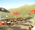 a terrace with tables and chairs, umbrellas, a red fence, and a view of the mountains