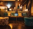 A cozy lounge area with a long bench, round tables, and two round chairs. The bench has multiple pillows, and the tables have potted plants on them. The room has wooden floors and a wooden ceiling with hanging lamps.