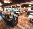 Wooden floor, wooden ceiling, large windows, buffet table, dining tables, chairs, plates, cups, toaster, bread, coffee machine, artwork on wall