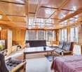 living room with wooden walls, sofa, coffee table, TV, lamp, bed, bedspread, carpet, wooden chair, wooden cabinet, shelves, books, cross, windows, curtains