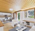 Open plan living room and kitchen, wooden ceiling, white walls, wooden floor, large windows, table, chairs, couch, coffee table, kitchen appliances, view of the outside