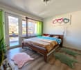 Bedroom with wooden bed, leopard print bedspread, two pink rugs, green rugs, green curtains, glass door leading to balcony, painting on wall, potted plant, wooden floor, ceiling lamp