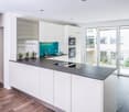 Modern open plan kitchen with island, white cabinets, black countertop, turquoise backsplash, glass door leading to balcony, built-in oven and microwave, stainless steel sink with faucet, ceramic floor.