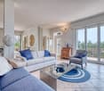 Spacious living room with large windows, gray sofa, blue armchairs, wooden dresser, and a round rug with a blue and white pattern on the tile floor. The room has access to a balcony or terrace.