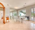 Spacious open-plan living area with tiled flooring, large windows, and a dining table with chairs. The room has an arched wooden doorway leading to another room.