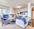 The image shows a spacious and well-furnished living room. The room features a large fireplace with a wooden mantle, which appears to be the focal point of the space. The living area is centered around a comfortable L-shaped sofa upholstered in a light-co