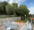 The image shows an outdoor patio area with stone walls, lounge chairs, potted plants, and a covered seating area. The patio is surrounded by lush greenery and trees, creating a peaceful and relaxing atmosphere.