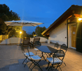 The image shows a cozy outdoor patio area of a residential property. The patio features a large umbrella-covered dining table with chairs, as well as a couple of lounge chairs. The patio is made of tiled flooring and is surrounded by lush greenery and tre