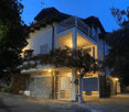 The image shows a multi-story residential building with a stone exterior and a balcony on the upper level. The building has a garage or covered parking area on the ground level. The property is surrounded by trees and landscaping, and the image is taken a