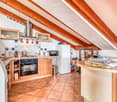 The image shows a cozy kitchen with a vaulted wooden ceiling and exposed beams. The kitchen features wooden cabinets, a tile backsplash, and a range oven. There is a refrigerator and a dishwasher visible. The floor is tiled, and there is a small dining ar