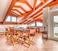 The image shows a spacious dining room with a high, exposed wooden beam ceiling. The room features a large wooden dining table surrounded by rustic wooden chairs. The room has a cozy and warm atmosphere, with the wooden beams and tile flooring adding to t