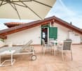 The image shows an outdoor patio area with a tiled floor and a large umbrella providing shade. The patio is furnished with a dining table and chairs, as well as lounge chairs. The exterior of the building features a wooden roof and green shutters, giving