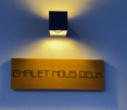 A wooden sign with the text 'CHALET NOUS DEUX' displayed on a blue wall, with a black square light fixture above it.